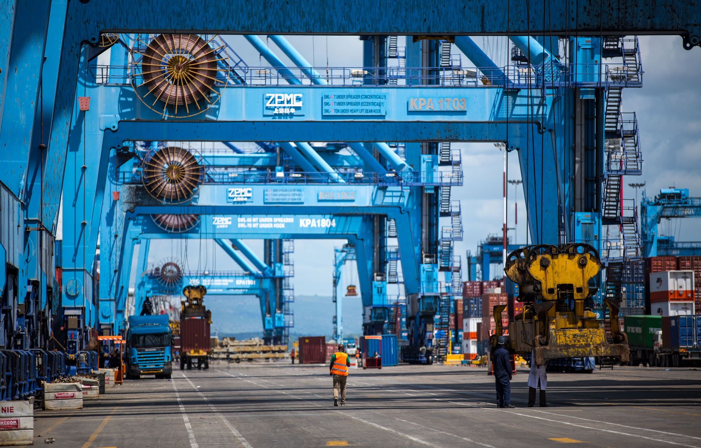Electrically powered ship-to-shore container cranes with large cable reels at the Port of Mombasa, with workers and trucks on the quay. The port is one of the industrial loads the project aims to serve; it is not the project site.